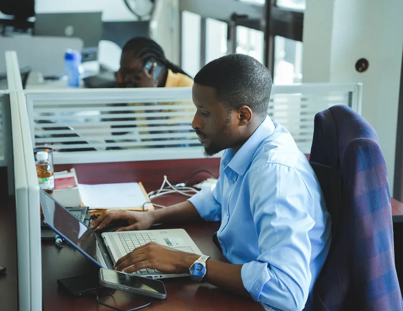 A man in a blue shirt working on a laptop at a desk in an office